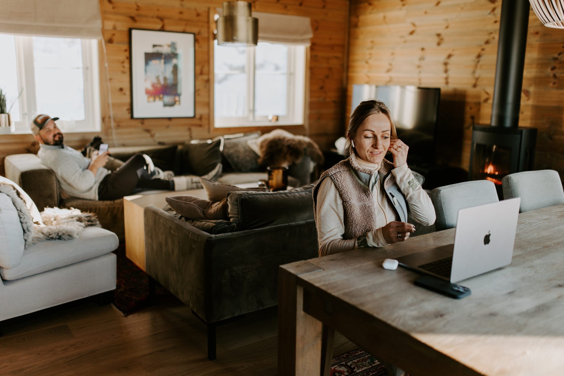 Par slapper av i koselig stue i hytten. Kvinne jobber på bærbar PC, mann hviler på sofaen. Varmt treinteriør med peis.