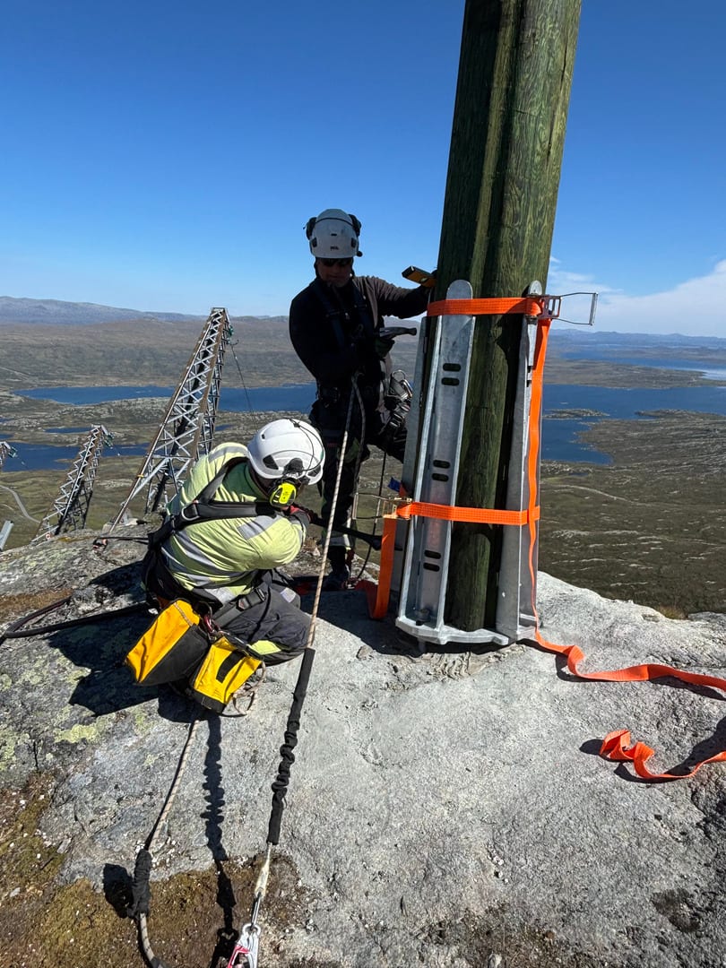 To linjearbeidere i verneutstyr jobber på toppen av et fjell med en ny trestolpe, sikret med tau og sele, med utsikt over innsjøer i bakgrunnen.