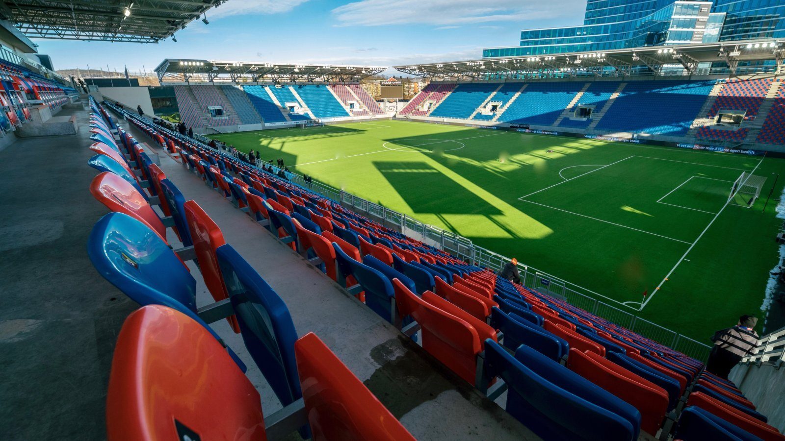 Empty stadium with red and blue seats overlooking a green football field and modern building.